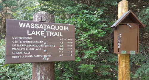 Trail sign for Wassataquoik Lake Trail with directions and distances to various points, surrounded by trees.