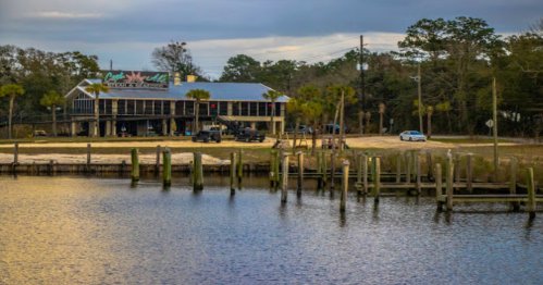 A waterfront restaurant with a deck, surrounded by palm trees and parked vehicles, under a cloudy sky.