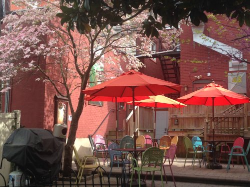 A cozy outdoor café with colorful chairs and red umbrellas, surrounded by blooming trees and a brick building.