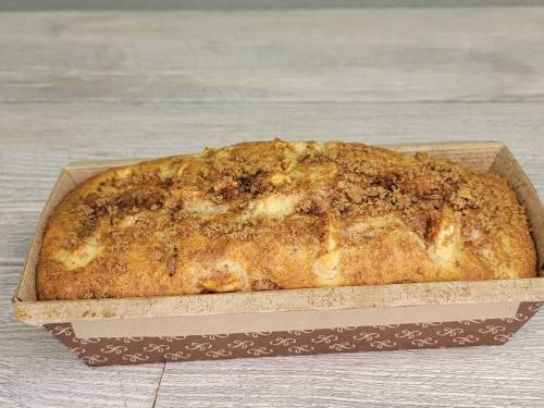 A freshly baked loaf of bread with a crumbly topping, placed in a brown paper baking dish on a wooden surface.