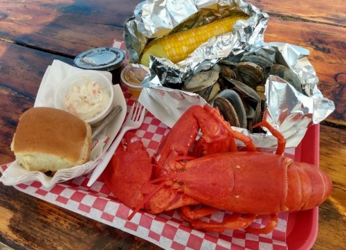 A seafood platter featuring a whole lobster, corn on the cob, clams, coleslaw, and a roll on a red-checkered tray.
