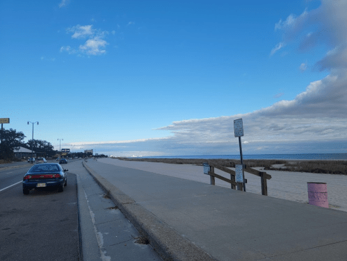 A coastal road with a parked car, leading to a sandy beach and a cloudy sky over the ocean in the distance.