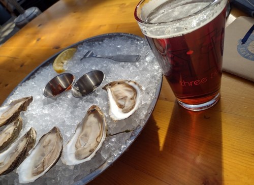 A plate of oysters on ice with a lemon wedge, next to a glass of amber beer on a wooden table.