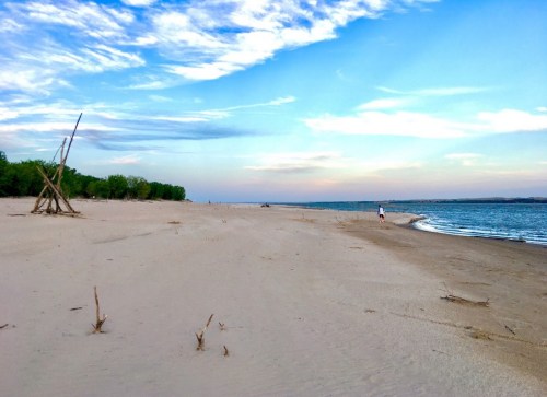 A serene beach scene with a sandy shore, gentle waves, and a person walking along the water under a blue sky.