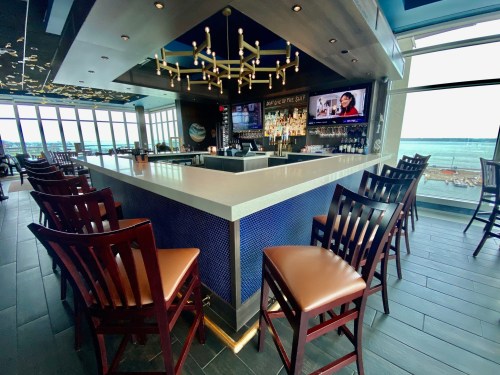 A modern bar with high stools, a sleek counter, and large windows overlooking a waterfront view.