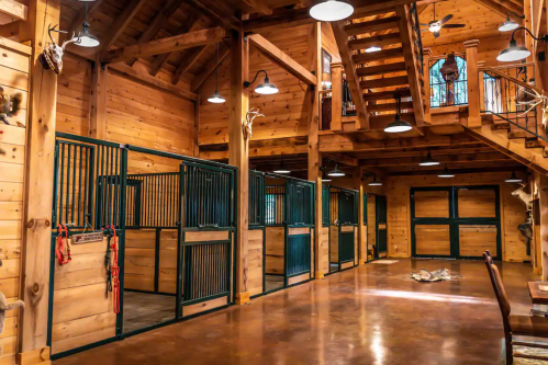 Interior of a wooden barn with horse stalls, a staircase, and rustic lighting.