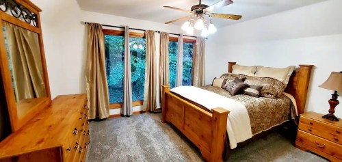 Cozy bedroom with a wooden bed, dresser, and large windows overlooking greenery. Soft lighting and neutral decor.