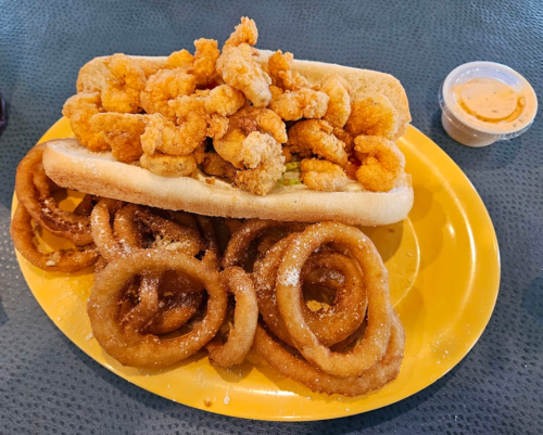 A plate featuring a shrimp po' boy sandwich topped with fried shrimp, alongside a serving of curly fries and dipping sauce.