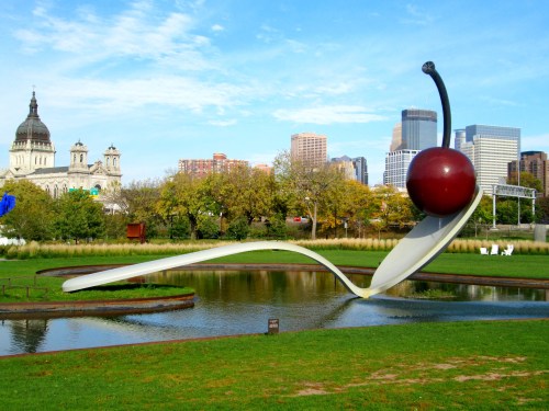 A large spoon with a cherry sculpture in a park, with city buildings and blue sky in the background.