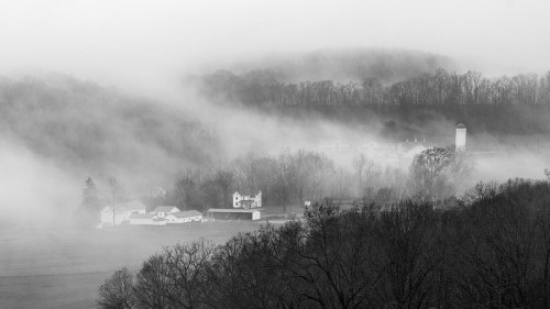 A misty landscape featuring a farmhouse and trees, shrouded in fog with rolling hills in the background.