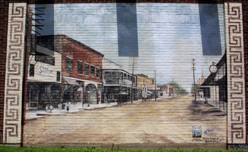 A mural depicting a historic street scene with buildings and a clear sky, painted on a brick wall.