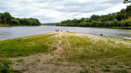 A sandy riverbank with green grass, people enjoying the area, and trees lining the opposite shore under a cloudy sky.
