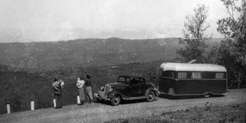 A vintage black-and-white photo of two people and a dog by a car and trailer, overlooking a scenic mountain view.