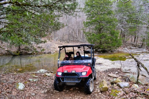 Two people sit in a red off-road vehicle near a stream, surrounded by trees and rocky terrain.