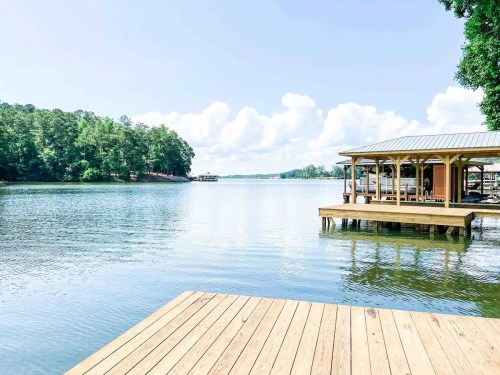 A serene lake view with wooden docks, surrounded by lush trees and a clear blue sky with fluffy clouds.