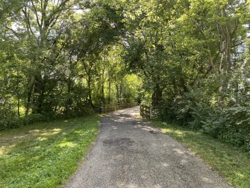 A gravel path surrounded by lush green trees, leading to a wooden fence in a serene, natural setting.