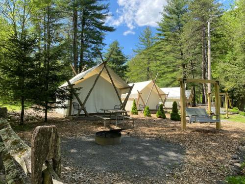 Two canvas tents in a wooded area, surrounded by trees, with a fire pit and a swing set nearby.