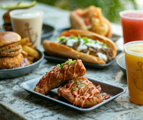 A close-up of crispy chicken wings drizzled with sauce, surrounded by burgers, hot dogs, and drinks on a table.