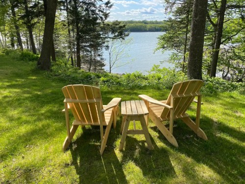 Two wooden Adirondack chairs and a small table sit on a grassy area by a serene lake, surrounded by trees.