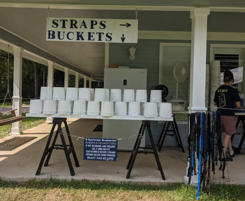 A booth with a sign for "Straps" and "Buckets," displaying white buckets and straps on stands. Staff member in the background.