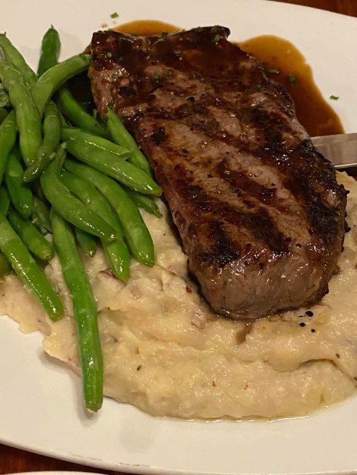 A grilled steak served with green beans and creamy mashed potatoes, drizzled with sauce on a white plate.