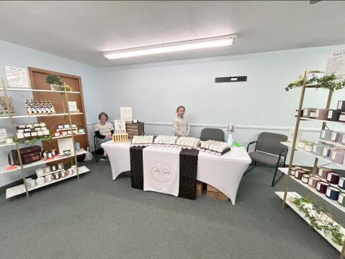 A small vendor booth with two women, displaying candles and skincare products on tables and shelves in a light blue room.
