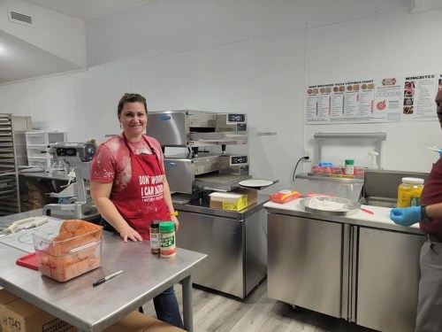 A smiling woman in an apron stands in a kitchen with cooking equipment and ingredients on the counter.