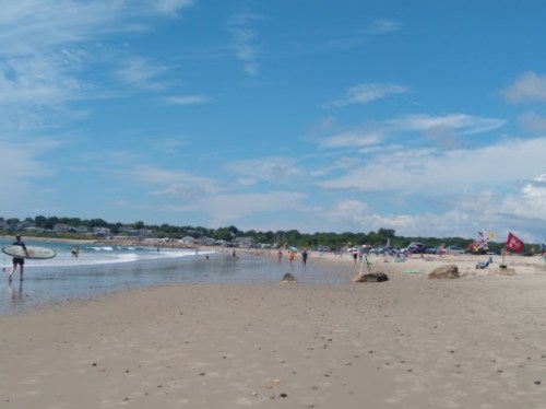 A sunny beach scene with people surfing, walking, and relaxing under colorful umbrellas along the shore.