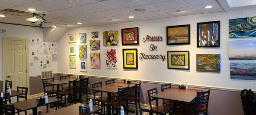 A cozy café interior featuring tables and a wall adorned with colorful artwork and the phrase "Artists In Recovery."