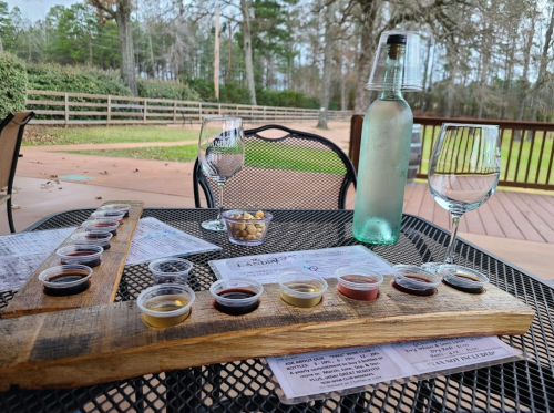A wooden flight board with various sauces, two wine glasses, a bottle of water, and snacks on a patio table.