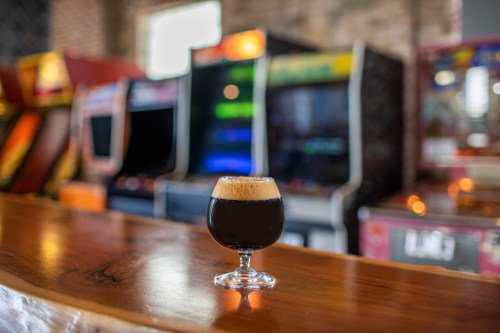 A glass of dark beer sits on a wooden bar, with vintage arcade machines blurred in the background.