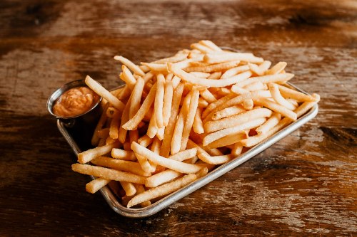 A metal tray filled with golden, crispy French fries and a small cup of dipping sauce on a wooden surface.
