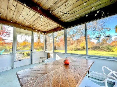 A cozy screened porch with a wooden table, surrounded by autumn foliage and string lights.
