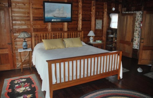 Cozy wooden bedroom with a large bed, yellow pillows, and rustic decor, featuring a ship painting and a colorful rug.