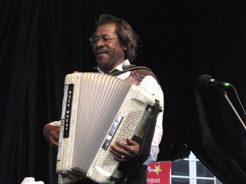 A musician playing an accordion on stage, wearing glasses and a white shirt, with a microphone nearby.
