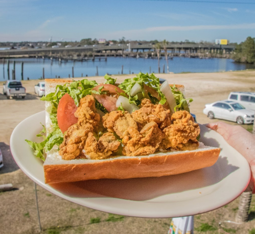 A hand holding a large sandwich filled with fried oysters, lettuce, tomato, and onions, with a scenic waterfront in the background.