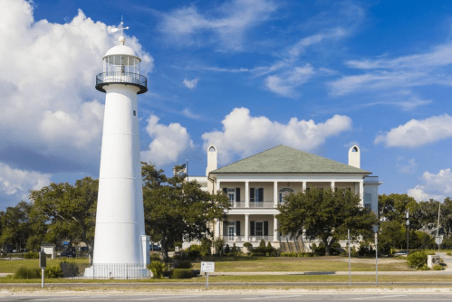 A white lighthouse beside a historic building with a green roof, set against a blue sky with fluffy clouds.