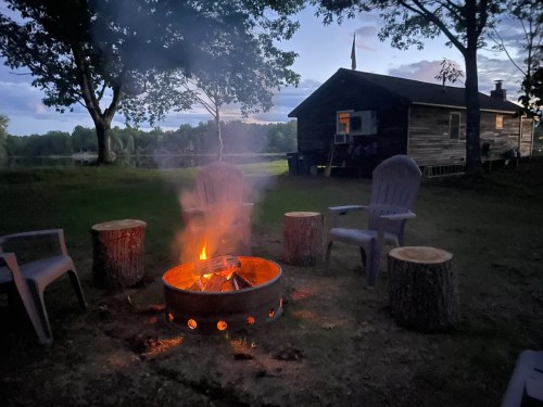 A cozy campfire surrounded by log seating, with a rustic cabin in the background under a twilight sky.