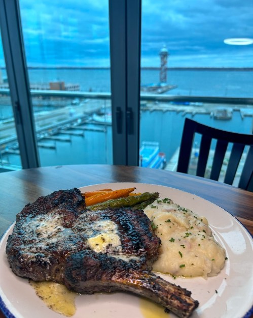 A beautifully plated steak with butter, mashed potatoes, and vegetables, overlooking a marina and cloudy sky.