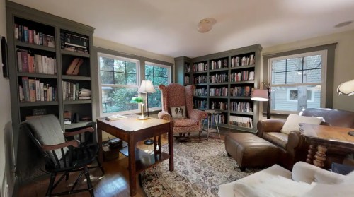 Cozy reading nook with bookshelves, a comfortable chair, a wooden table, and large windows letting in natural light.
