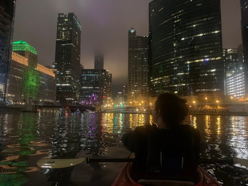A person in a kayak on a city river at night, surrounded by illuminated skyscrapers and a foggy skyline.
