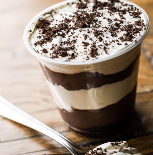 A layered dessert in a clear cup, featuring chocolate and cream topped with crushed cookies, on a wooden surface.