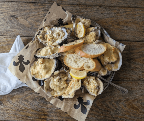 A plate of baked oysters topped with breadcrumbs, lemon slices, and served with slices of bread on a wooden table.