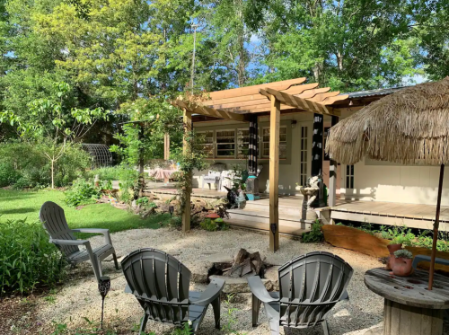 A cozy outdoor space with a wooden deck, seating area, and lush greenery, featuring a fire pit and thatched roof structure.