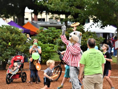 A costumed character throws a frisbee to children at a lively outdoor event with tents and families enjoying the day.