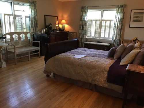 Cozy bedroom with a large bed, wooden furniture, and natural light from windows. A seating area is visible in the corner.