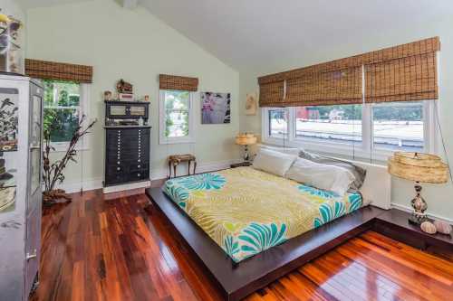 Bright bedroom with a wooden floor, large windows, and a bed featuring a colorful floral blanket.