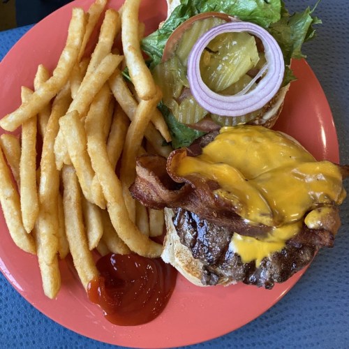 A plate with a cheeseburger topped with bacon, lettuce, tomato, pickles, and onion, served with a side of fries and ketchup.