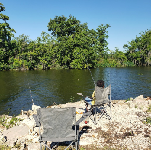 Two fishing chairs by a riverbank, with a person sitting in one chair, surrounded by lush greenery and clear blue skies.