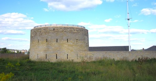 A round stone structure with a flat roof, surrounded by grass and under a blue sky with scattered clouds.